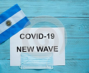 On a blue wooden background is a sheet with the inscription: covid -19 new wave, a medical mask and the flag of Argentina