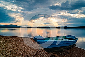 Blue wooden boat moored on the shore of lake under cloudy sky at sunset