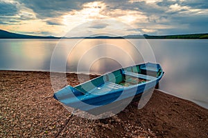 Blue wooden boat moored on the shore of lake under cloudy sky at sunset