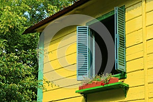 Blue wooden window shutters on vivid yellow house among green foliage in sunlight