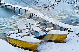 Blue and yellow catamaran in a winter day, covered with snow standing near the frozen river and wooden bridge.