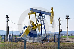 Blue and yellow pump jack above an oil well surrounded by electricity poles and protected with a fence, in bright daylight.