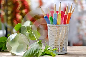 blue, yellow, red colored pencils with lead, glass ball globe, green plant branch on wooden table, writing supplies, concept Back