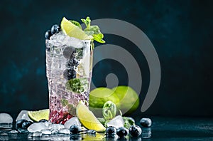 Blueberry Mojito cocktail drink with lime, white rum, soda, cane sugar, mint, and ice in glass on deep blue background. Summer