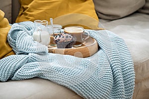 Blueberry muffin with coffee, sugar and milk on wooden tray and comfy sofa with blue wool knit blanket and yellow cushions