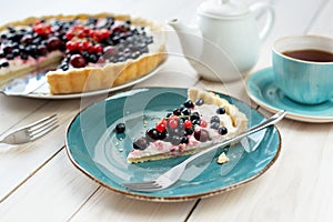 Blueberry open pie on blue plate with cup of black tea and white tea pot on bright painted wood background