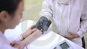Blurred Asian doctor putting on blood pressure gauge on arm of unrecognizable pregnant Caucasian woman. Professional