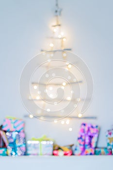 Blurred background of Unusual Christmas tree made of wooden sticks with festive lights on white wall and colourful gift boxes.