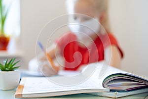 Blurred blonde schoolgirl in medical mask studying at home, doing school homework, writing in notebook. Reading training photo