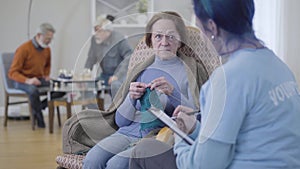 Blurred Caucasian volunteer talking with female retiree in nursing home. Elderly woman knitting and gesturing no by