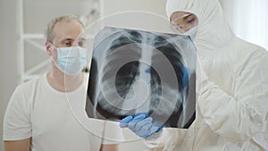 Blurred doctor in antiviral suit talking to mid-adult man in face mask showing Covid-19 complications on lungs roentgen