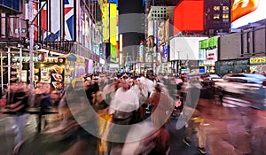 Blurred faces in the crowds of people walking down 7th Avenue past the bright colorful lights of Times Square in New York City at