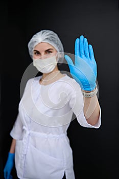 Blurred female healthcare worker in white lab coat and medical mask, shows her hand palm to camera. STOP sign gesture