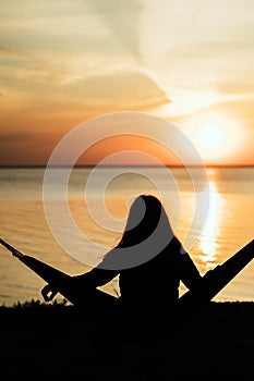 Blurred girl sitting on a hammock in a pine forest on the edge of a cliff near the sea and watching the sunrise