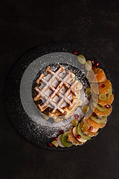 Blurred image of Belgian waffles with powdered sugar and fruits on a dark background, top view