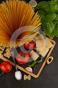 Blurred image of different types of pasta, tomato, basil, seasoning on a dark background