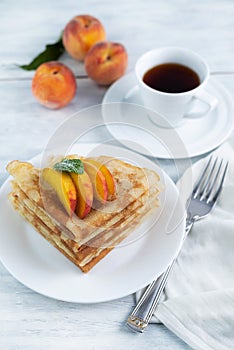 Blurred image of pancakes with pieces of peach in a plate, fork and a cup of tea on a light table