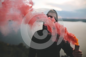 Blurred image of Ultras hooligan holding red smoke bomb in hand, standing on top of rock mountain with amazing view on river.