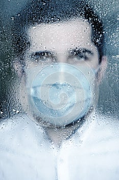 Blurred image of young man in protective mask standing behind the window with rain drops.