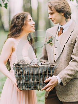 The blurred lovely smiling newlyweds is holding woven box with gray ferrets in the forest.