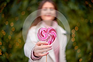 Blurred lovely woman extending red heart-shape lolly-pop on stick to camera. Focus on candy on forefront