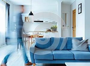 A blurred man walking in a modern apartment interior with a blue sofa and wooden kitchen cabinets, long exposure
