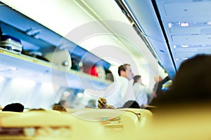 Blurred photo of airplane interior, stewards and passengers with bags on shelves.