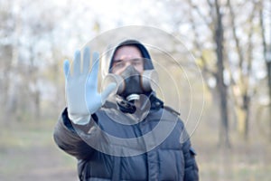 Blurred portrait of young man in protective gas mask and rubber disposable gloves shows stop gesture outdoors in spring wood