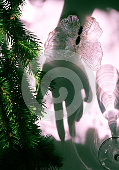 Blurred shadow of female hand and branch of Christmas tree, christmas butterfly toy and wine glass with light passes through a