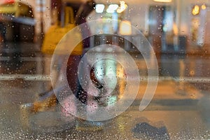 Blurred silhouette of an abstract unrecognizable girl with a handbag, view through a wet window with raindrops, city