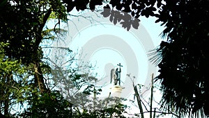 Blurred silhouette statue of an angel with a sword against the blue sky in the frame of foliage