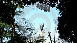 Blurred silhouette statue of an angel with a sword against the blue sky in the frame of foliage