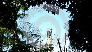 Blurred silhouette statue of an angel with a sword against the blue sky in the frame of foliage