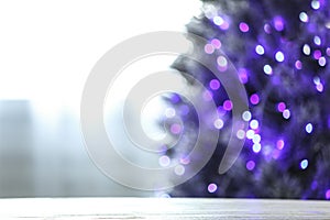Blurred view of beautiful Christmas tree with purple lights near window indoors, focus on wooden table. Space for text