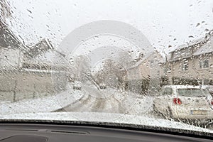 Blurred view of snow covered street through wet windshield of car on snowy, rainy day. Water drops from melting snow on window