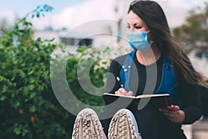 Blurred young woman in a medical mask in a park with a notebook and pen, writes.