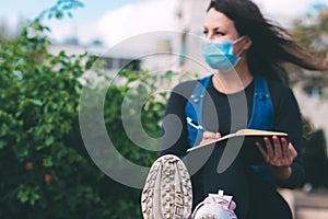 Blurred young woman in a medical mask in a park with a notebook and pen, writes.