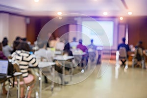 Blurry of auditorium for shareholders` meeting or seminar event with projector and white screen, many business people listening o