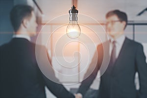 Blurry businessmen shaking hands together on abstract office interior background with glowing light bulb. Work ideas and