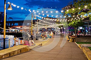 Blurry people hanging out at waterfront boardwalk string lights, beach chairs, patio restaurant dining tables sunset blue hour