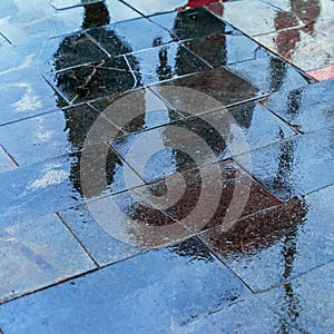 Blurry reflection shadows silhouettes of a woman under umbrella and a man walking in the rain in the city sidewalk