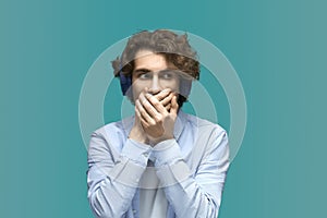 Blurt out. Portrait of a young beautiful man wearing white t-shirt and blue shirt in headphones closes his mouth with his hands