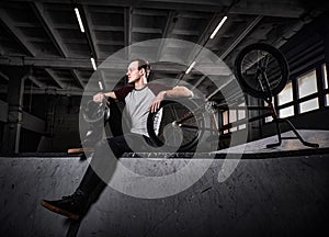 Bmx rider relaxing after practicing tricks with his bike in a skatepark indoors