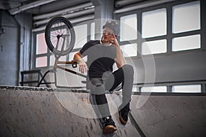 Bmx rider relaxing after practicing tricks with his bike in a skatepark indoors