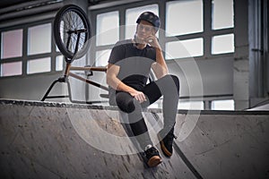 Bmx rider relaxing after practicing tricks with his bike in a skatepark indoors