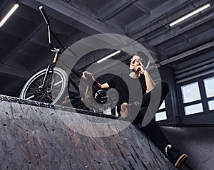 Bmx rider relaxing after practicing tricks with his bike in a skatepark indoors