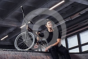 Bmx rider relaxing after practicing tricks with his bike in a skatepark indoors