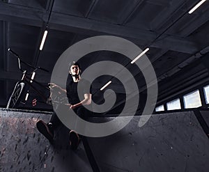 Bmx rider relaxing after practicing tricks with his bike in a skatepark indoors