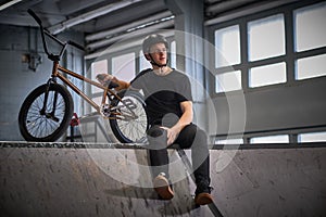 Bmx rider relaxing after practicing tricks with his bike in a skatepark indoors