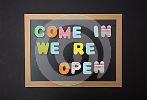 Board with black frame, text Come in, we re open in colorful letters, black wall background